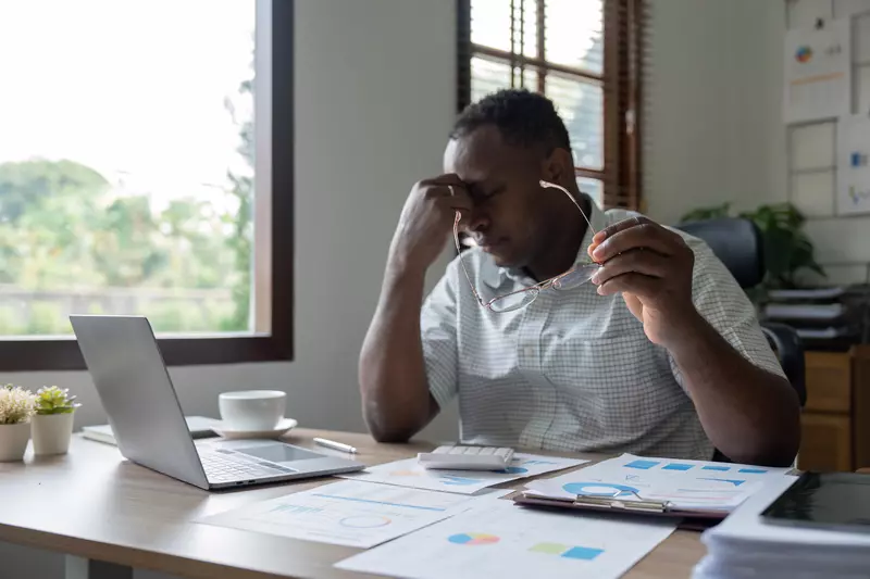 Man holding his forehead in frustration while looking down at a pile of documents next to his laptop
